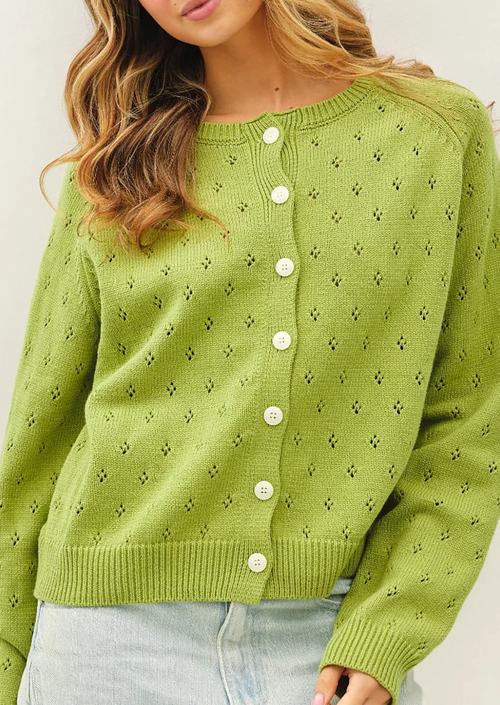 A woman wearing a light green knit cardigan sweater with white buttons, paired with light blue jeans, shown from the chest down against a plain light background.