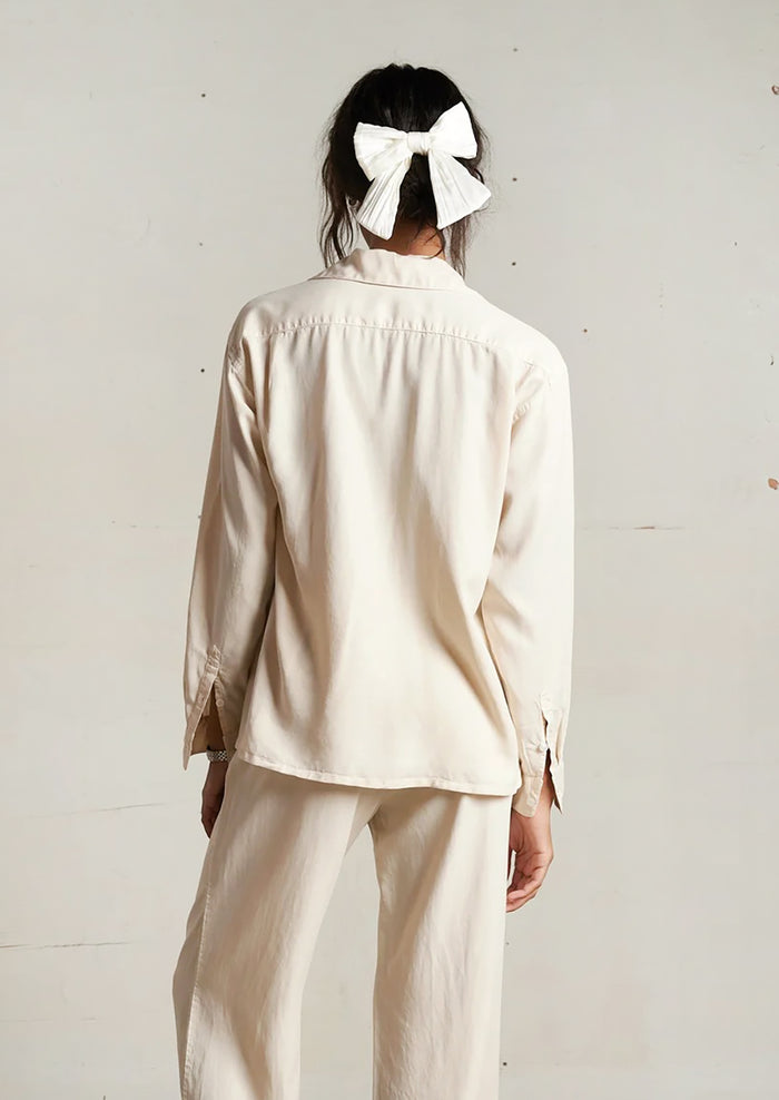 Person wearing a beige outfit with a white bow in their hair against a plain background