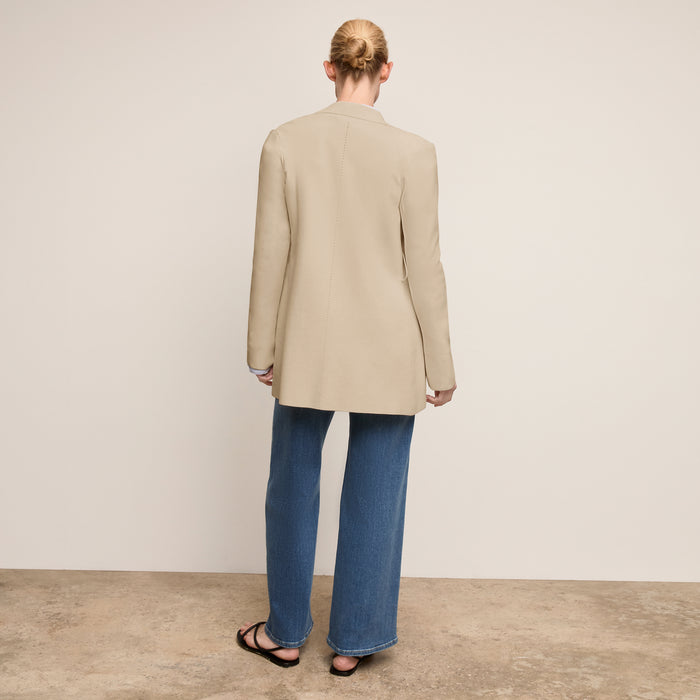 Person wearing a beige blazer and blue jeans standing against a plain background